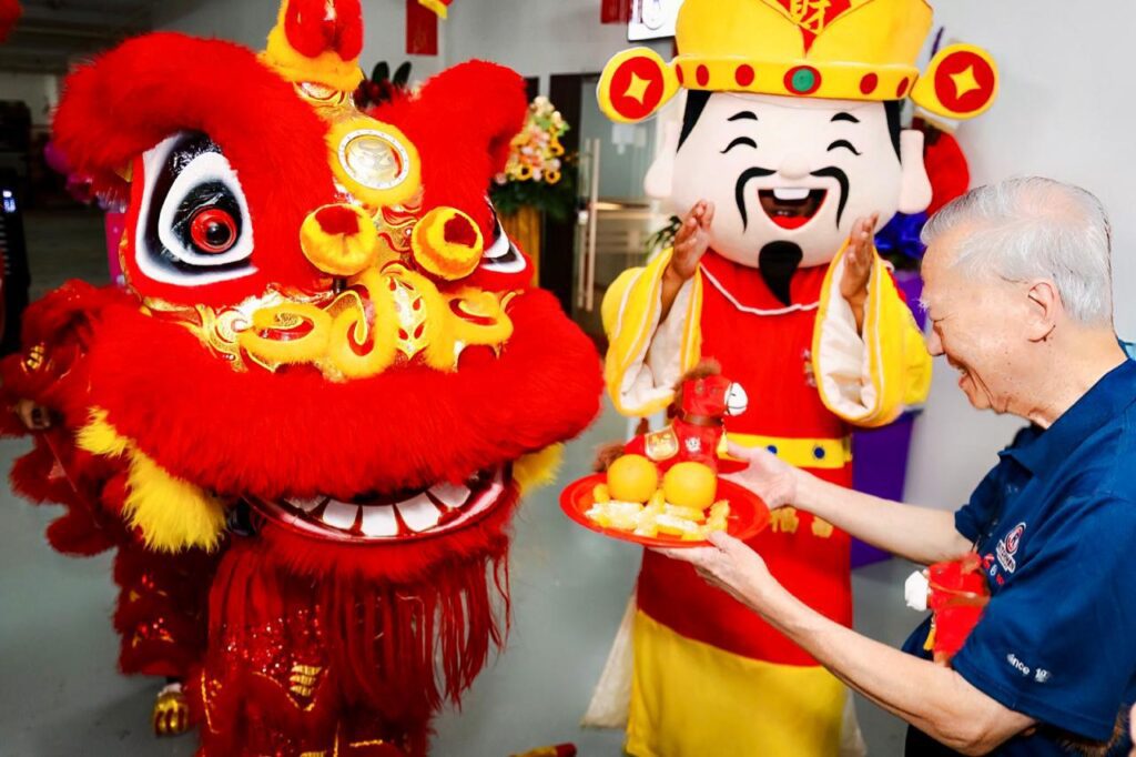 Ritchmen founder Mr C.M. Quek offering mandarin oranges to a lion dance lion, with a God of Fortune character behind.