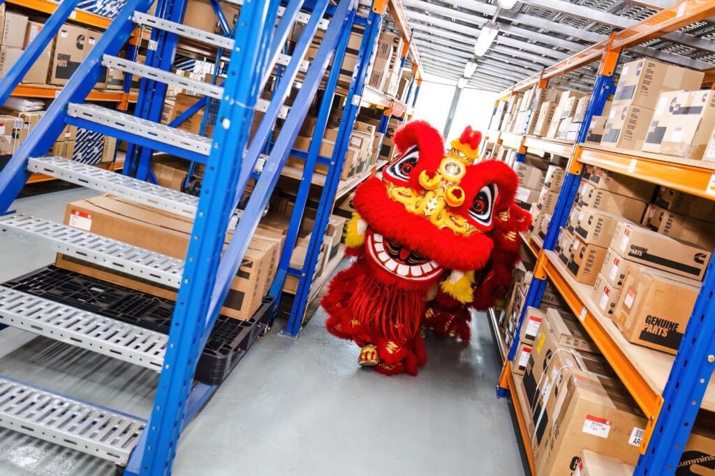A lion dance performer moving down an aisle of Cummins Genuine Parts cartons in the Ritchmen warehouse.